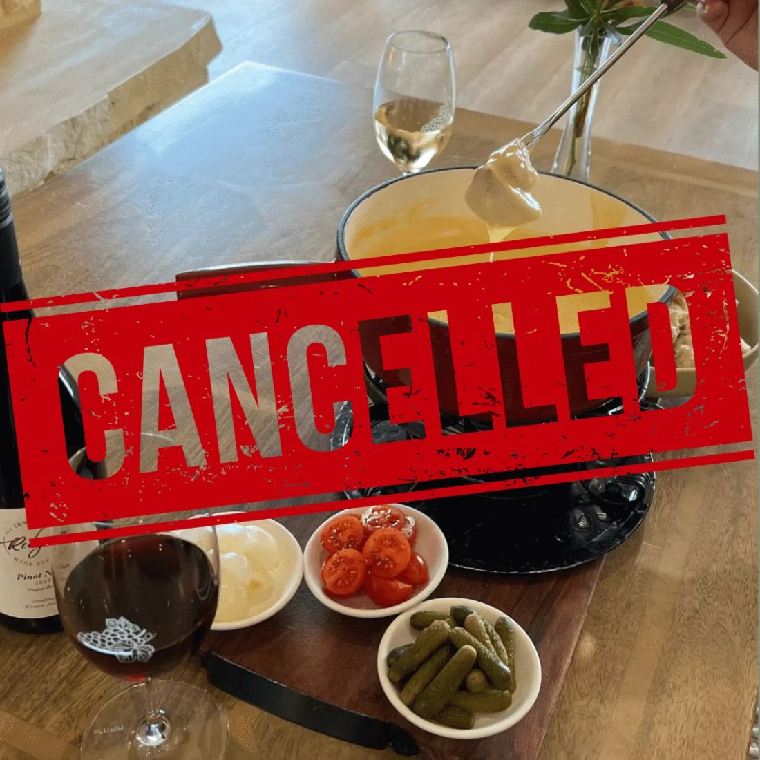 Cheese fondue pot with dipping items including bread, cherry tomatoes, pickles, and onions, accompanied by a bottle of red wine and glasses of red and white wine on the side, with a “Cancelled” badge overlaid on the image.