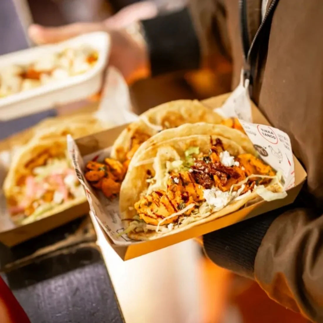 A person holds a tray with two soft tacos filled with grilled meat, shredded lettuce, cheese, and other toppings. Another food tray is visible in the background.