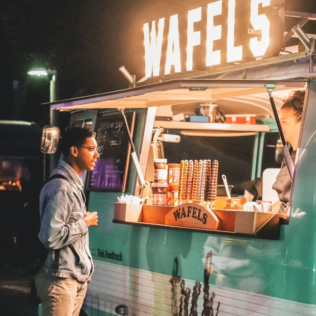 A person stands at a turquoise food truck with a bright WAFELS sign, looking at various waffle sticks displayed on the counter at night.