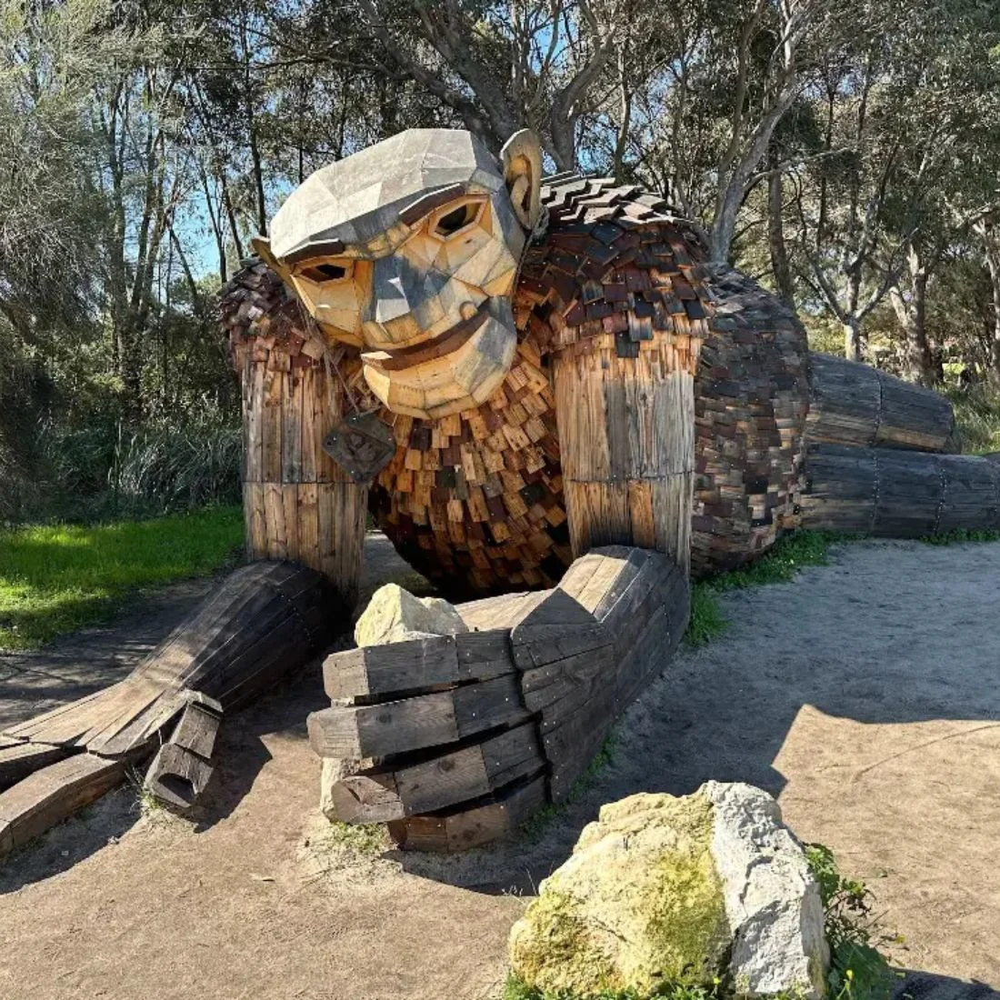A large wooden sculpture of a reclining troll with a human-like face is set outdoors among trees and grass, made from various pieces of wood in different shades.