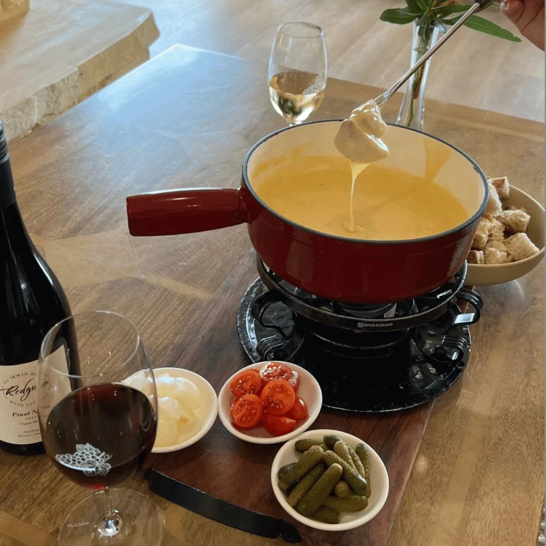 A cheese fondue pot with a piece of bread being dipped into it, accompanied by a bottle of red wine, a glass of red wine, a glass of white wine, and small bowls of cherry tomatoes, pickles, and onions.