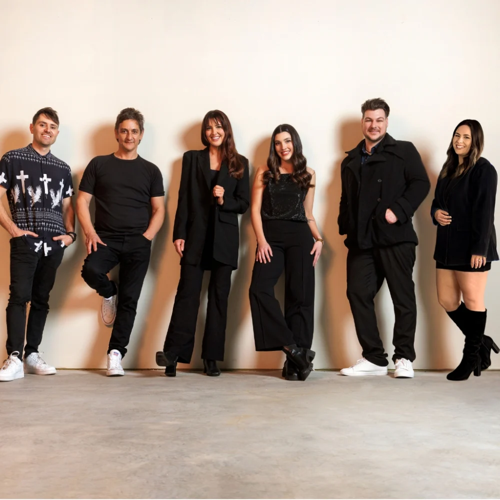 Six people stand in a row against a plain light-colored wall, smiling at the camera. They are dressed in mostly black attire, with varied outfits and relaxed poses.