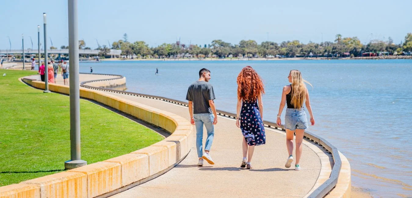 Three people walk along a curved waterfront path on a sunny day. The path is bordered by green grass on one side and blue water on the other.