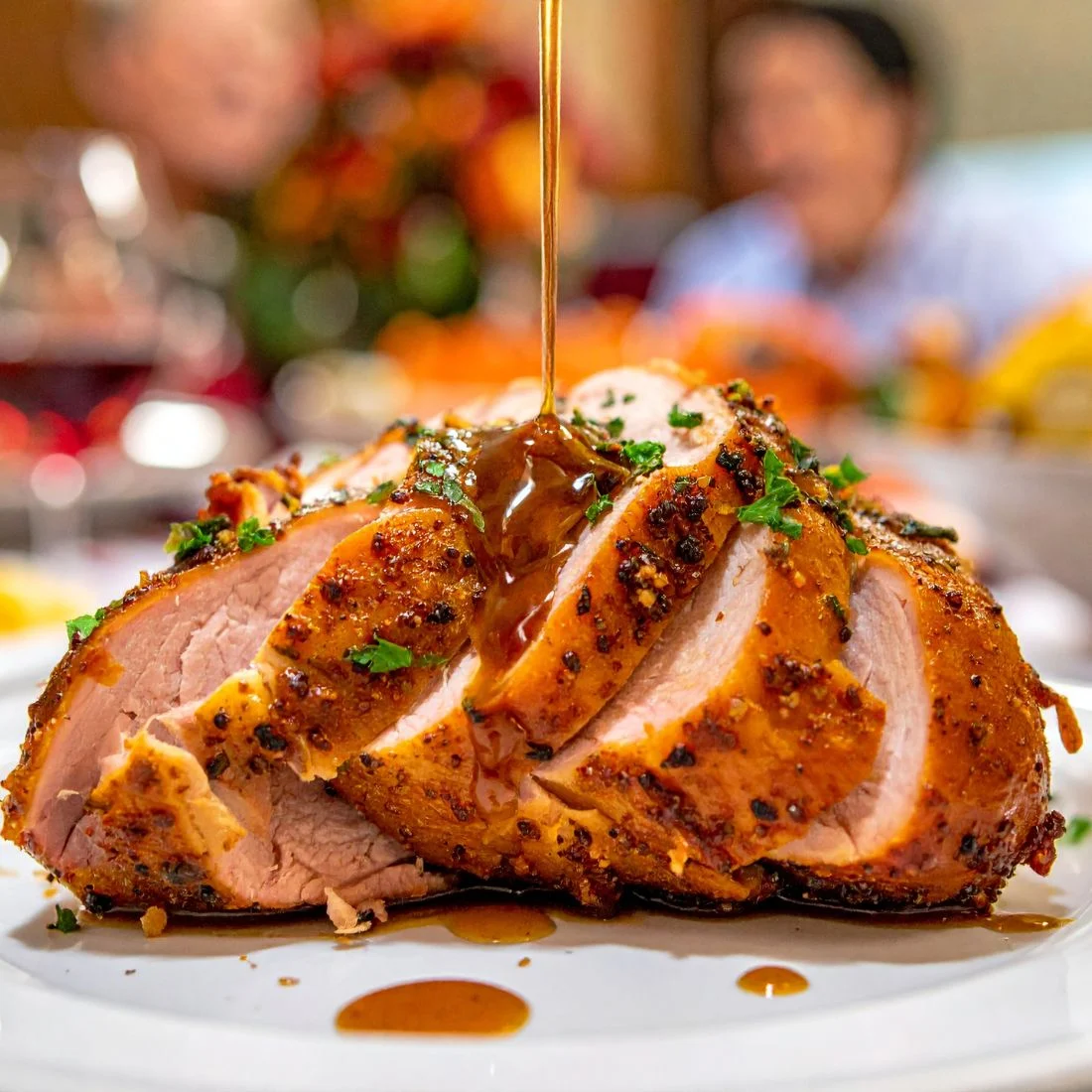 A sliced roasted turkey breast, seasoned with herbs and spices, is drizzled with gravy on a white plate. The dish is garnished with chopped parsley and appears to be part of a festive meal.