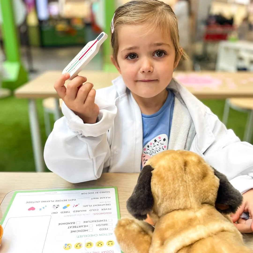 A young child in a white coat holds up a toy thermometer, sitting at a table with a stuffed dog and a medical chart, pretending to be a veterinarian.