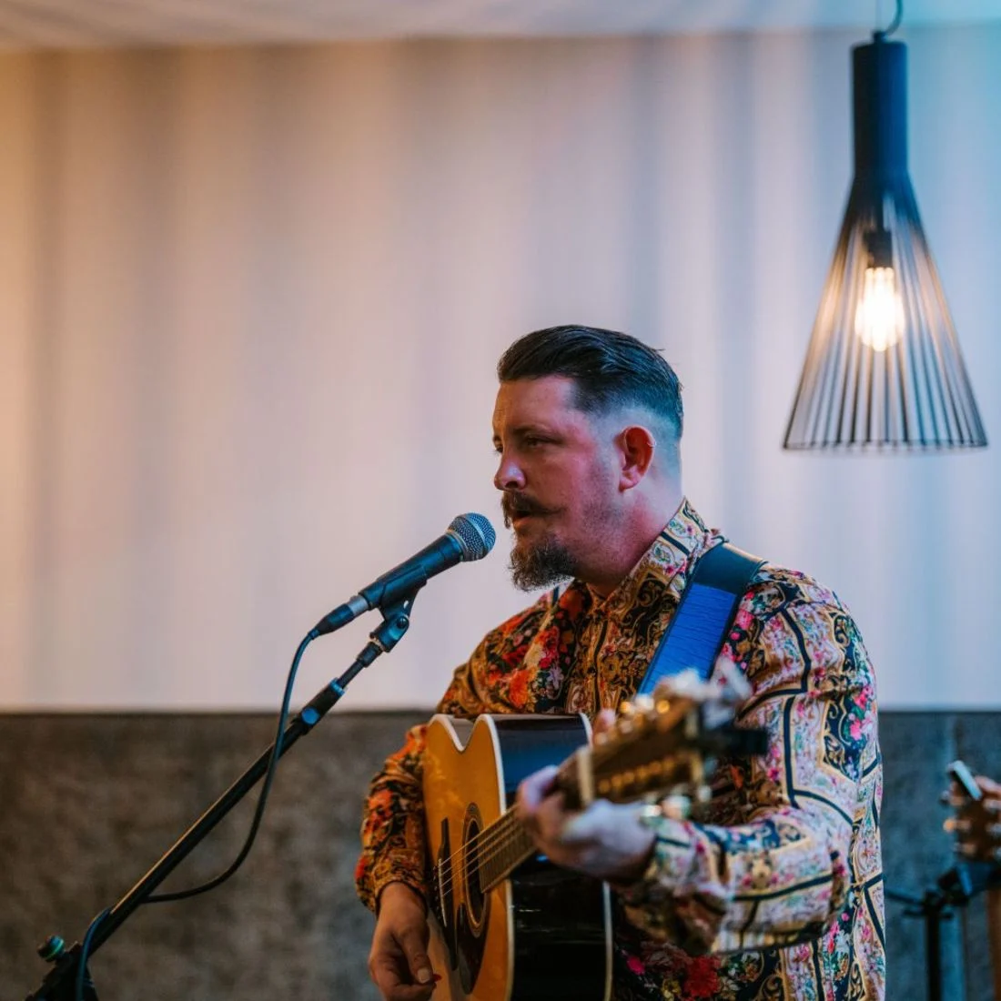 A man with a mustache and beard plays an acoustic guitar and sings into a microphone. He wears a colorful patterned shirt and a blue guitar strap. A modern light fixture hangs from the ceiling behind him.