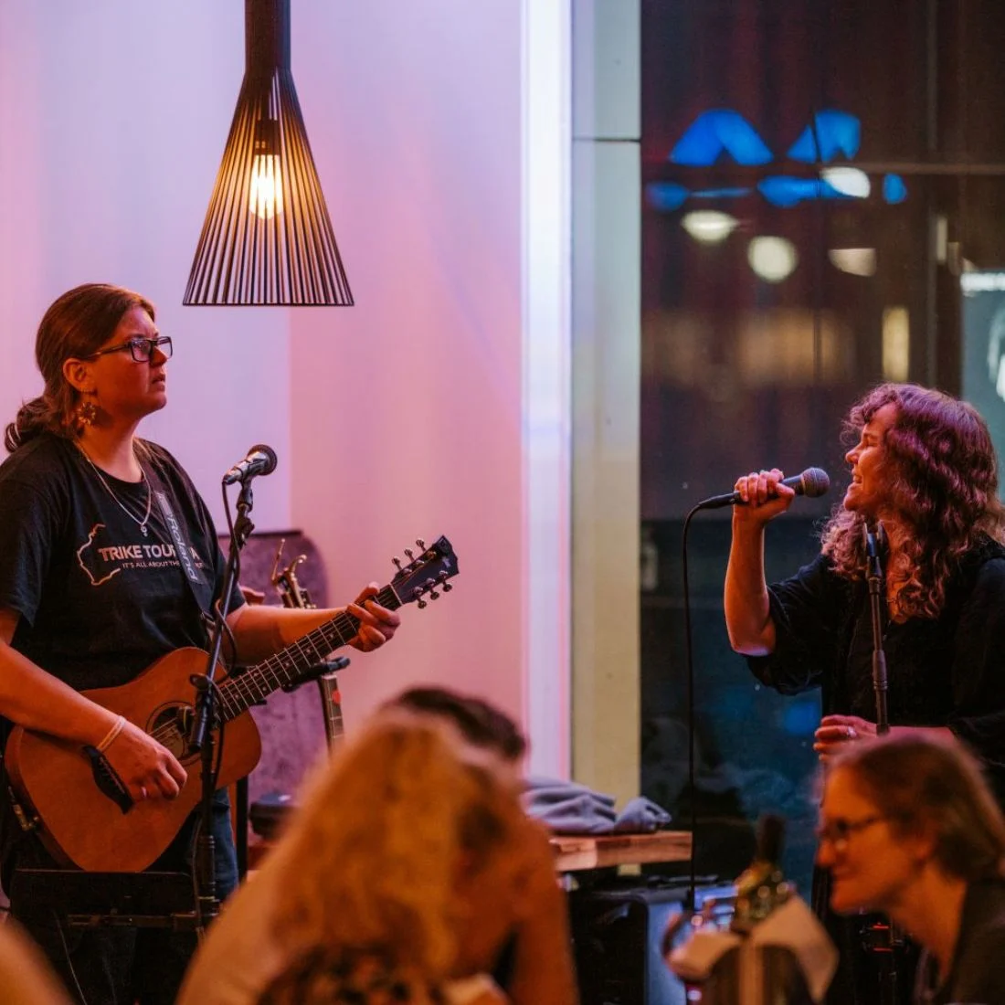 A woman plays an acoustic guitar and sings into a microphone while another woman sings into a microphone. They are performing indoors with warm lighting.