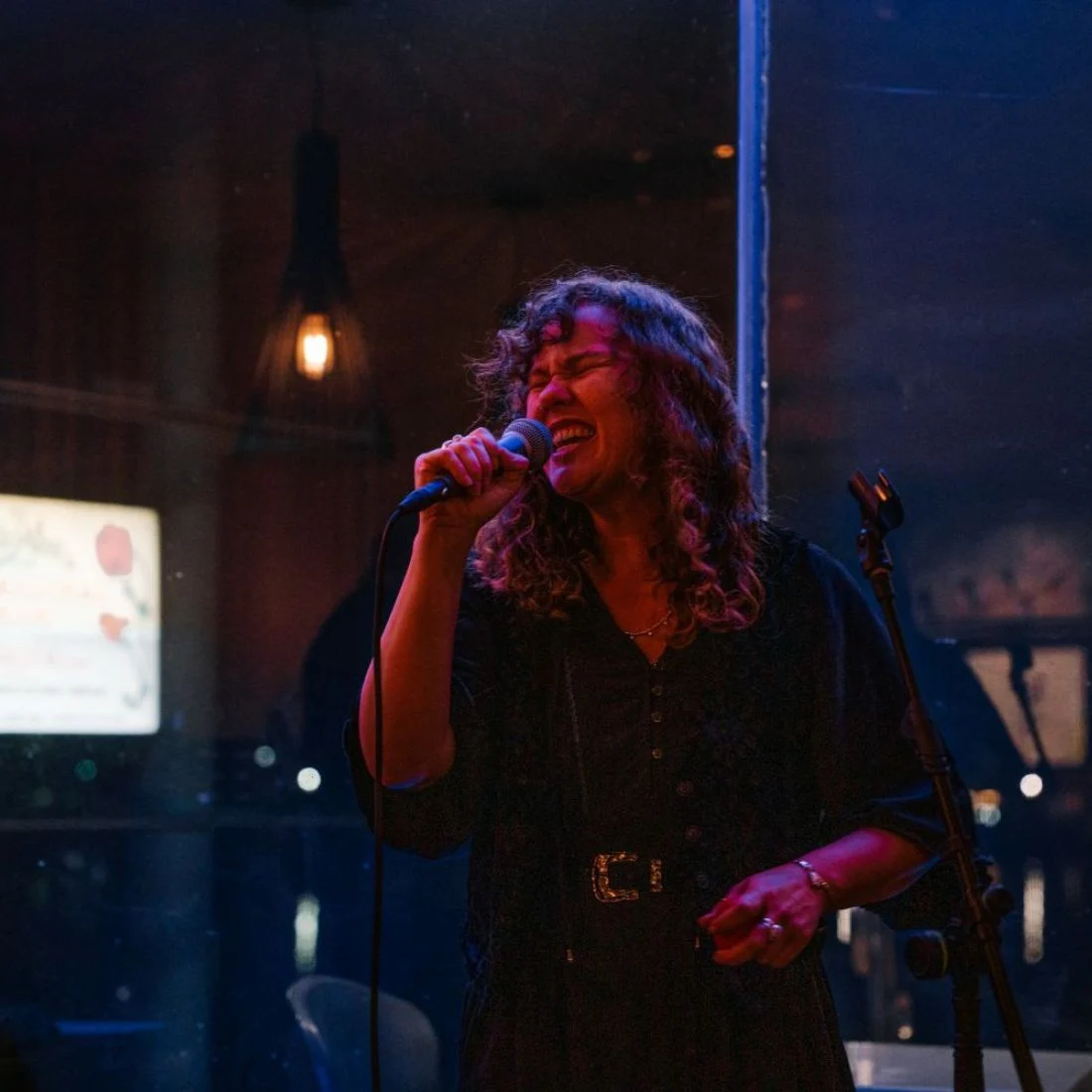 A woman with curly hair sings into a microphone on a dimly lit stage, wearing a dark dress with a distinctive belt.