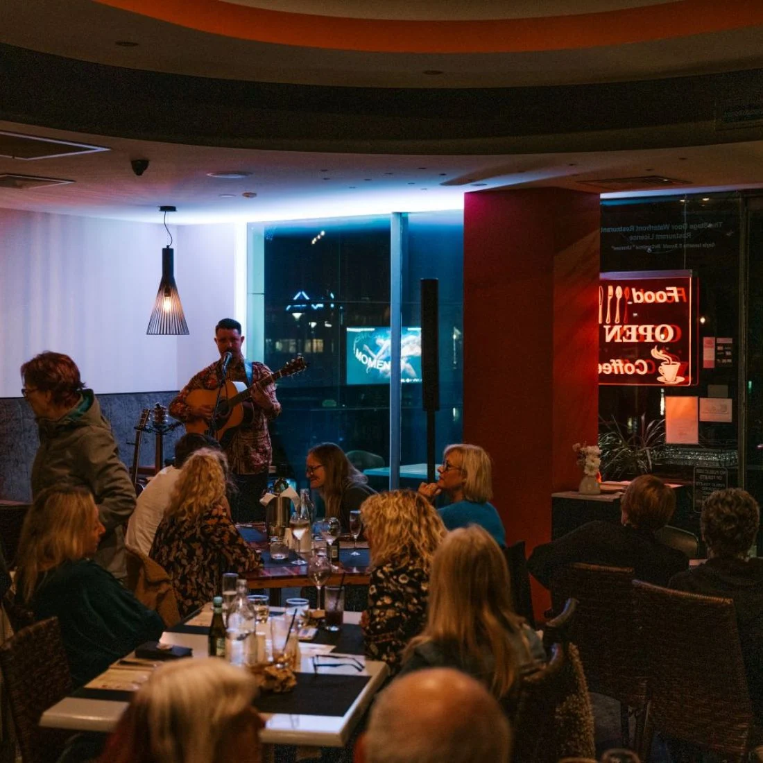 A man plays guitar and sings for an audience seated at tables in a dimly lit restaurant. A neon sign in the window reads 'Food Open Coffee'.
