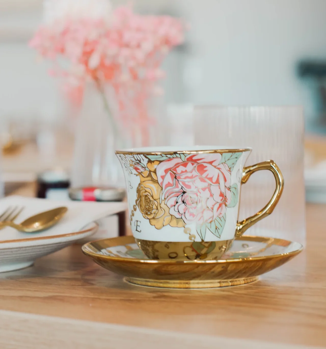 A decorative floral teacup with a gold handle and saucer sits on a wooden table, with blurred pink flowers and glassware in the background.