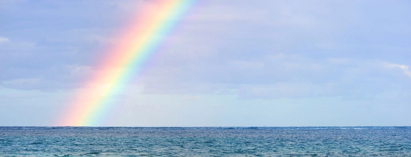 A rainbow arcs across a blue sky with clouds over a calm ocean.
