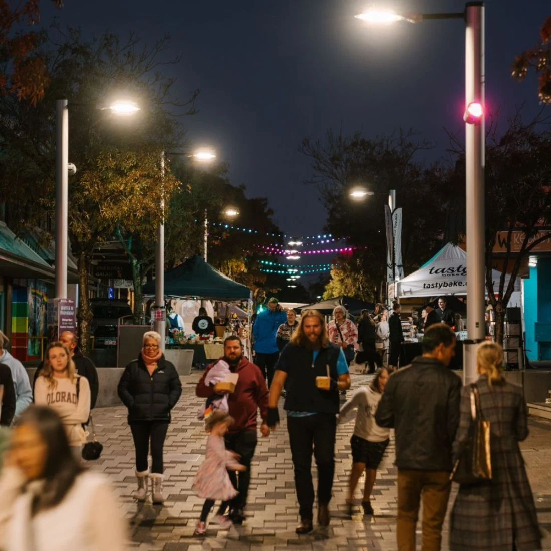 A street market at dusk with people browsing stalls, illuminated by overhead lights and colorful string lights.