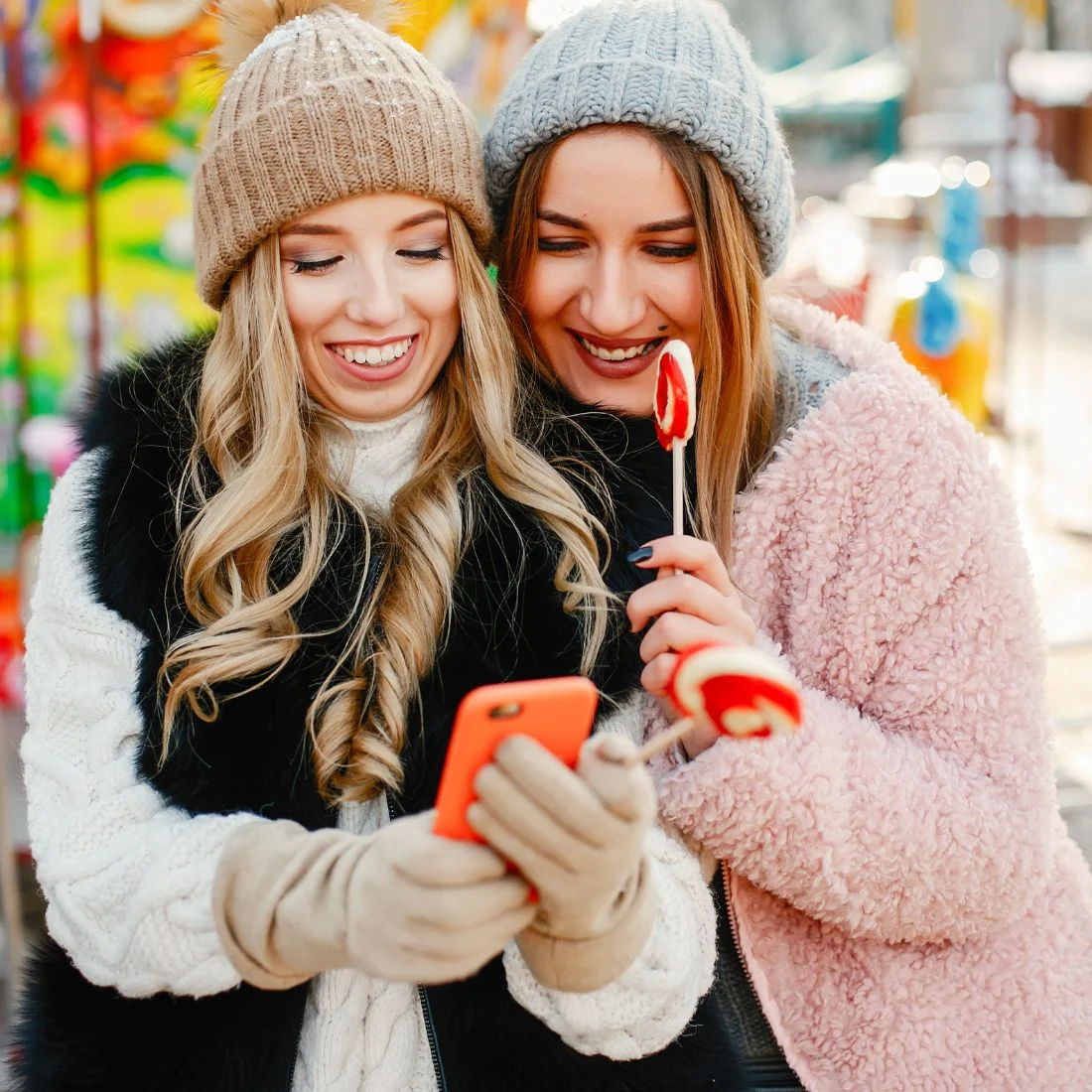 Two smiling young women wearing winter hats and coats look at a smartphone while holding lollipops.