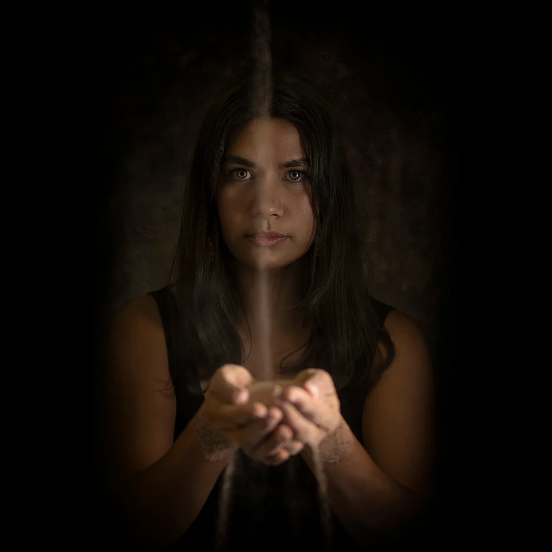 A woman with long dark hair holds out her hands as sand pours into them from above, surrounded by darkness, with only her face and hands illuminated.