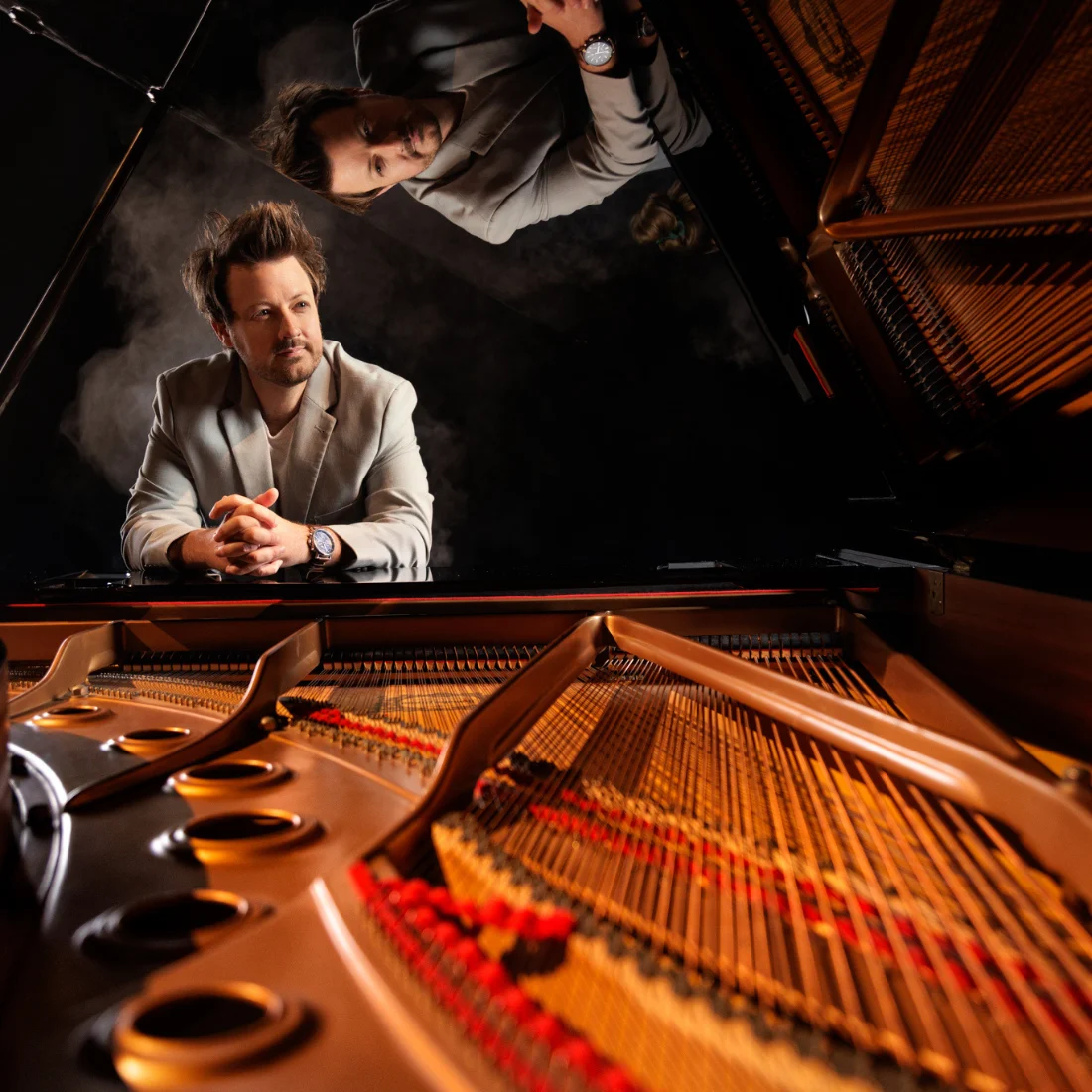 A man in a light suit sits at a grand piano, viewed from inside the piano, with strings and hammers visible. His reflection appears on the piano’s lid above him.
