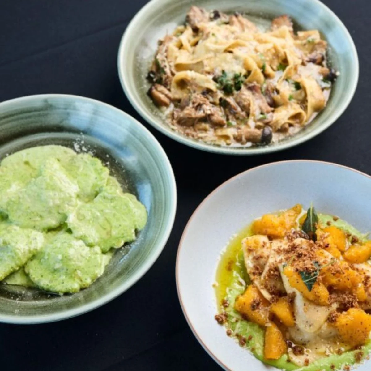 Three bowls of pasta dishes: one with green sauce and ravioli, one with tagliatelle and mushrooms, and one with fish, orange slices, and a green puree.