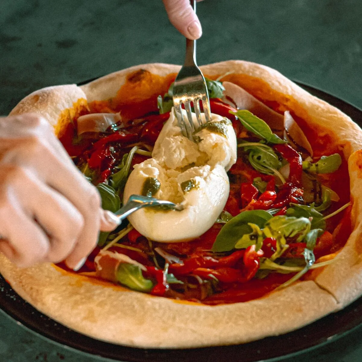 A person cuts into burrata cheese on a pizza topped with arugula, red peppers, and sauce.