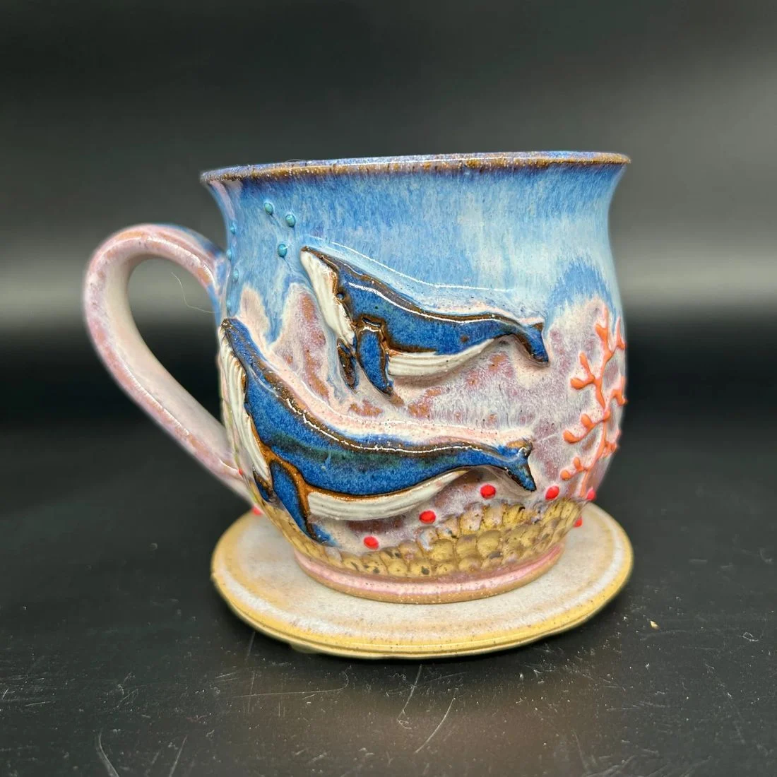 A ceramic mug with a matching saucer, decorated with detailed, raised designs of two blue whales swimming among coral and bubbles on a light blue and pink background.