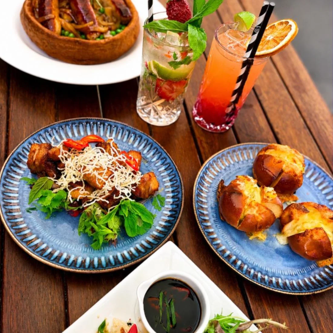Two blue plates with savory dishes, two colorful cocktails, cheesy bread rolls, and a bowl of stew are arranged on a wooden table for a vibrant meal.