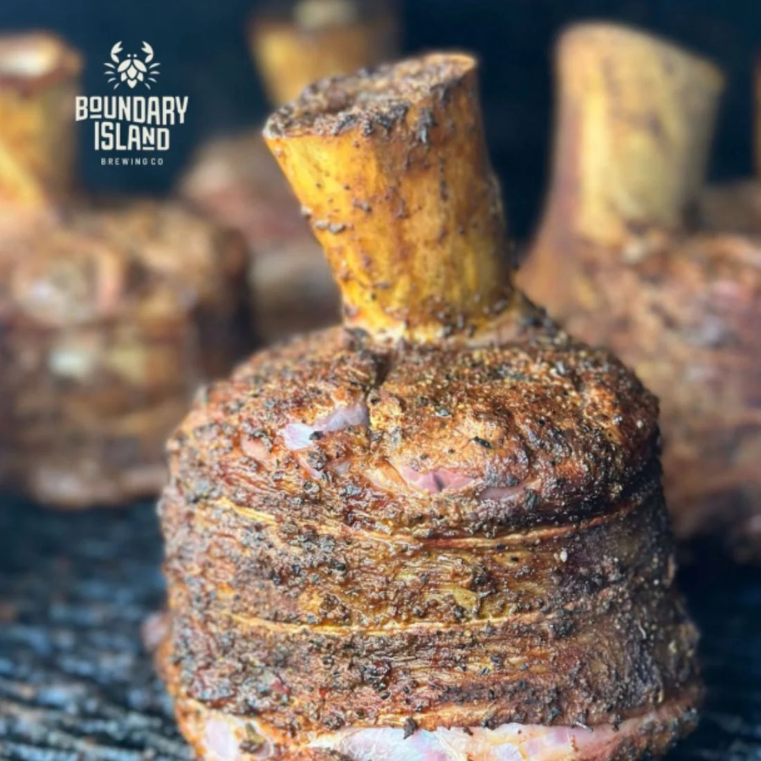 Close-up of several seasoned and grilled meat shanks on a grill, with the Boundary Island Brewing Co. logo in the top left corner.