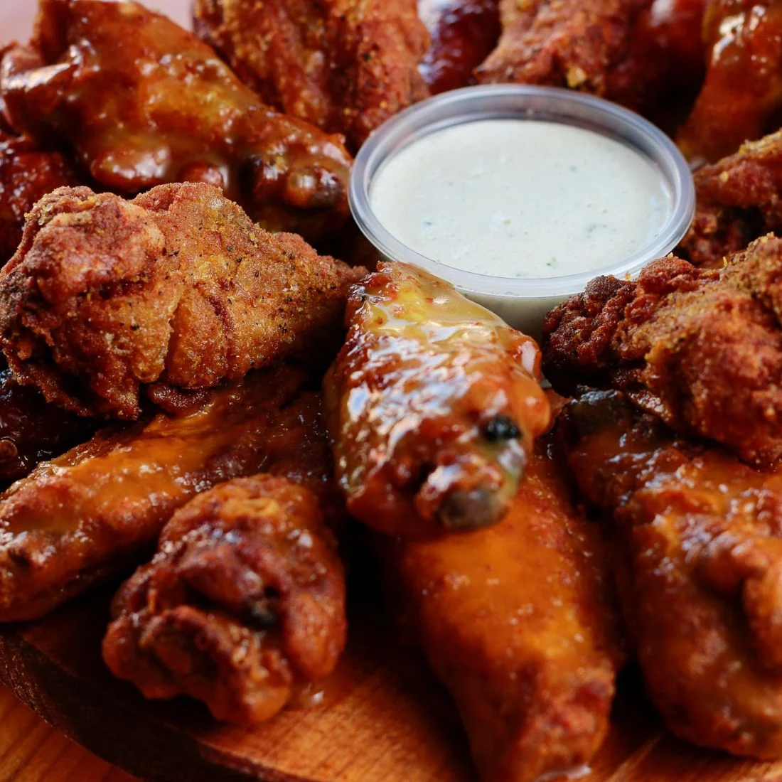 A close-up of sauced and fried chicken wings arranged around a plastic cup of ranch dipping sauce on a wooden plate.