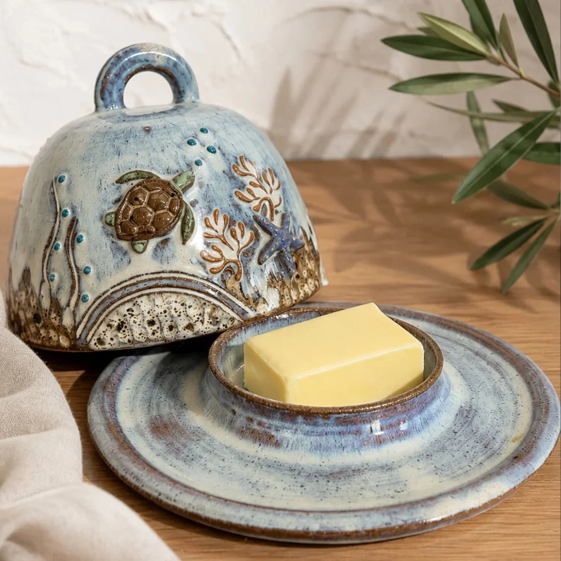 A ceramic butter dish with a sea turtle and ocean design sits on a wooden surface, holding a rectangular piece of butter. A leafy branch and beige cloth are nearby.