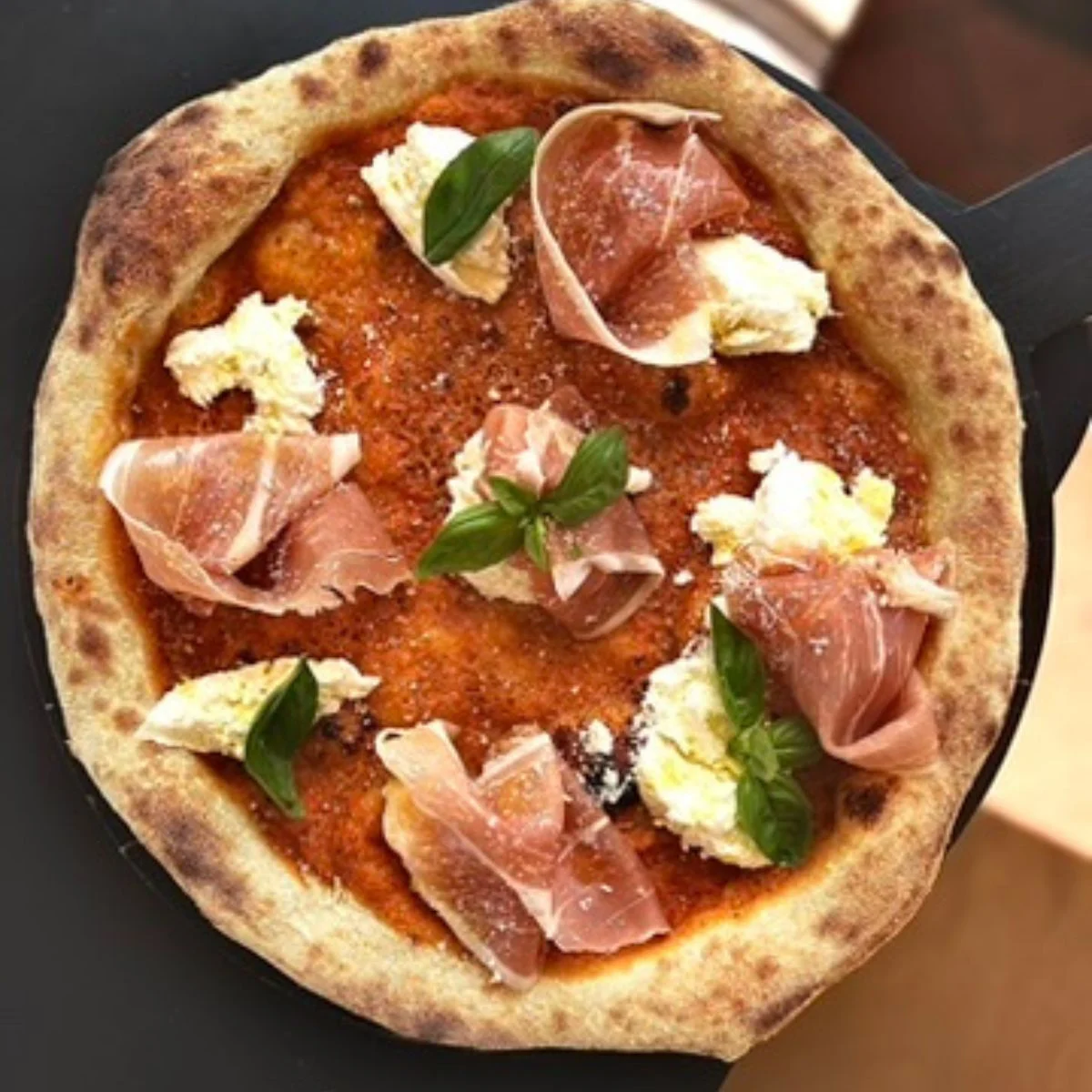 A round pizza topped with tomato sauce, dollops of ricotta cheese, prosciutto slices, and fresh basil leaves.