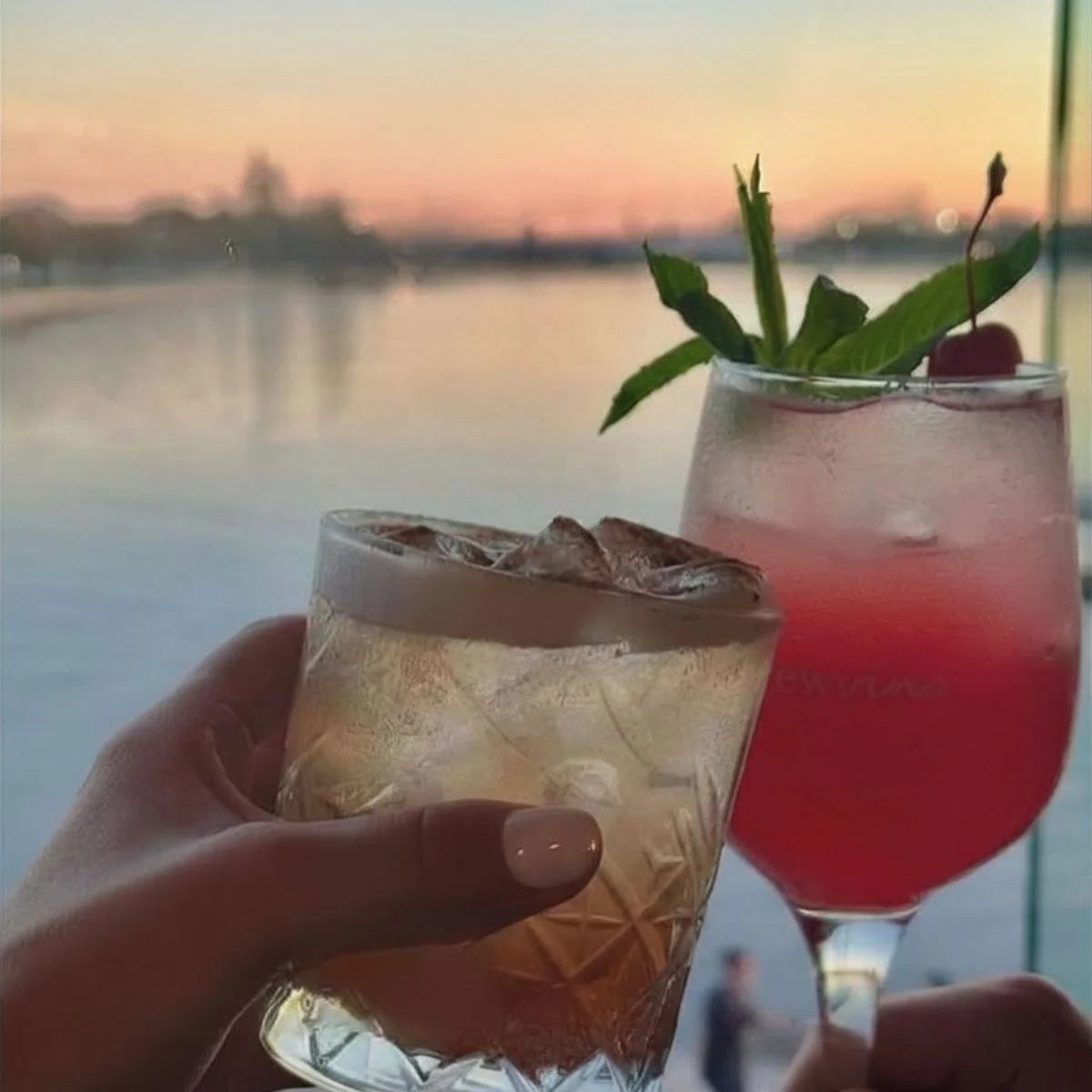 Two cocktails, one amber-colored with foam and ice, the other pink with mint and a cherry, are held up against a sunset over water.