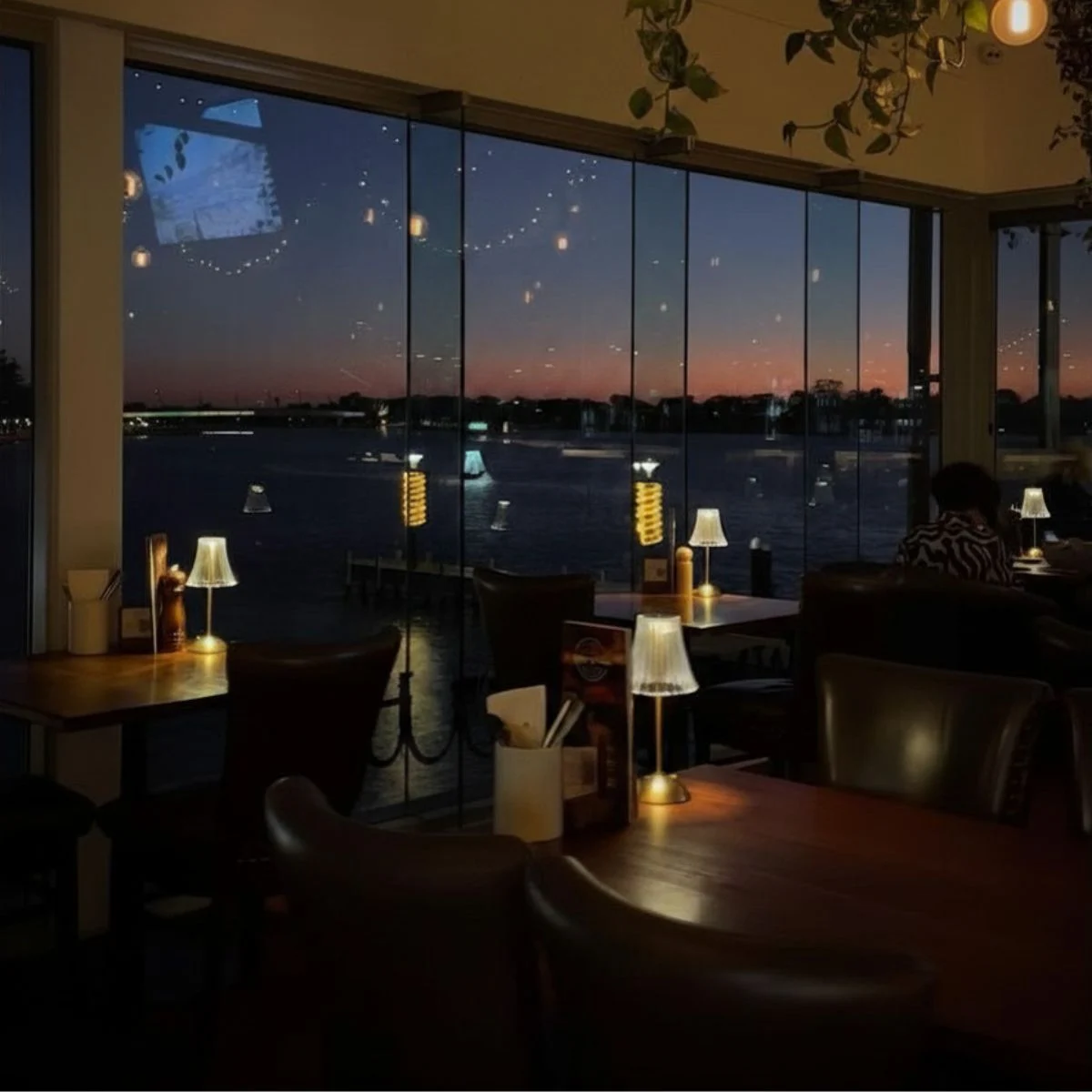 A dimly lit restaurant with large windows overlooking a body of water at dusk, with string lights and table lamps creating a warm ambiance.