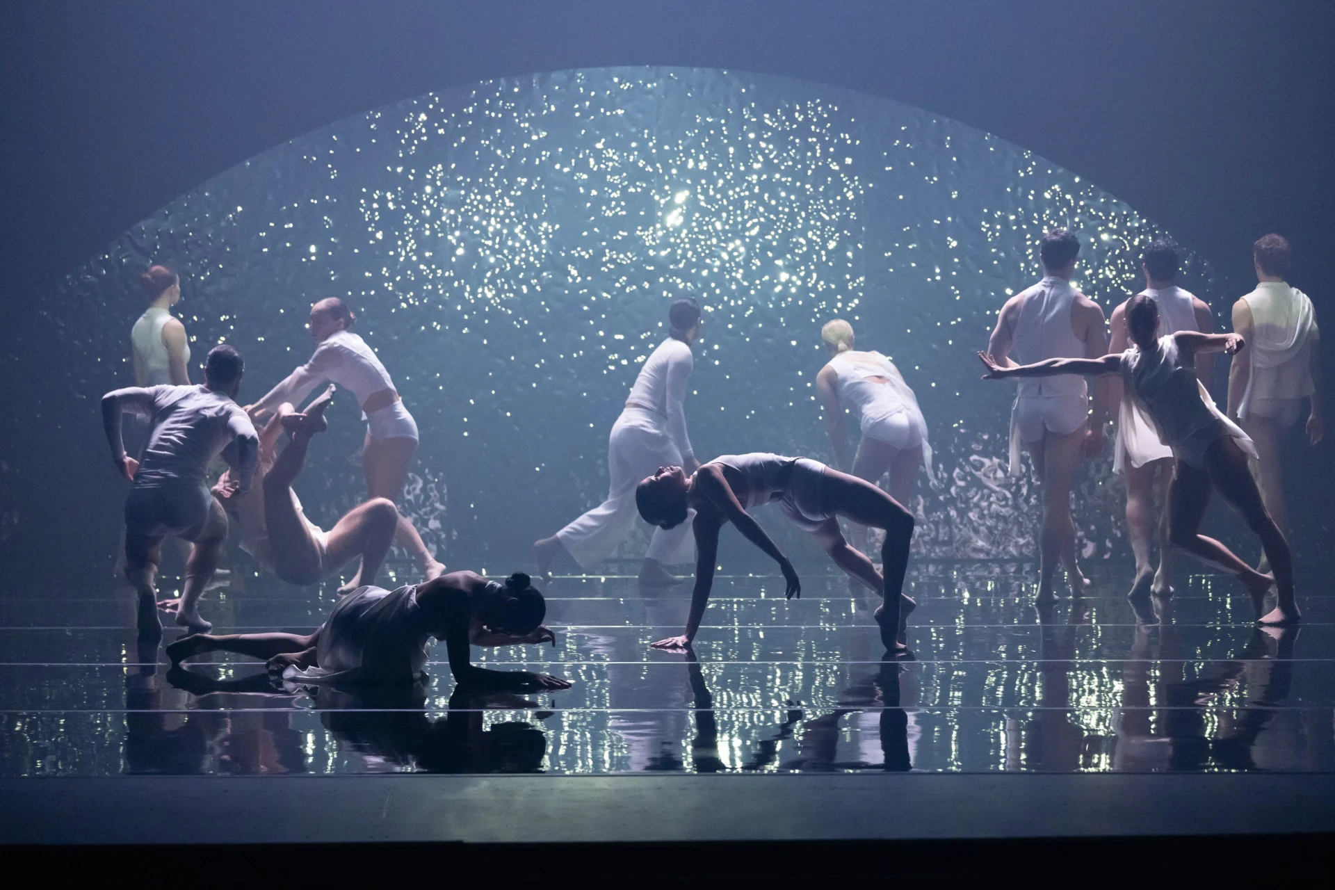 A group of dancers in white costumes perform on a reflective stage, with a sparkling, illuminated backdrop creating a dramatic and ethereal atmosphere.
