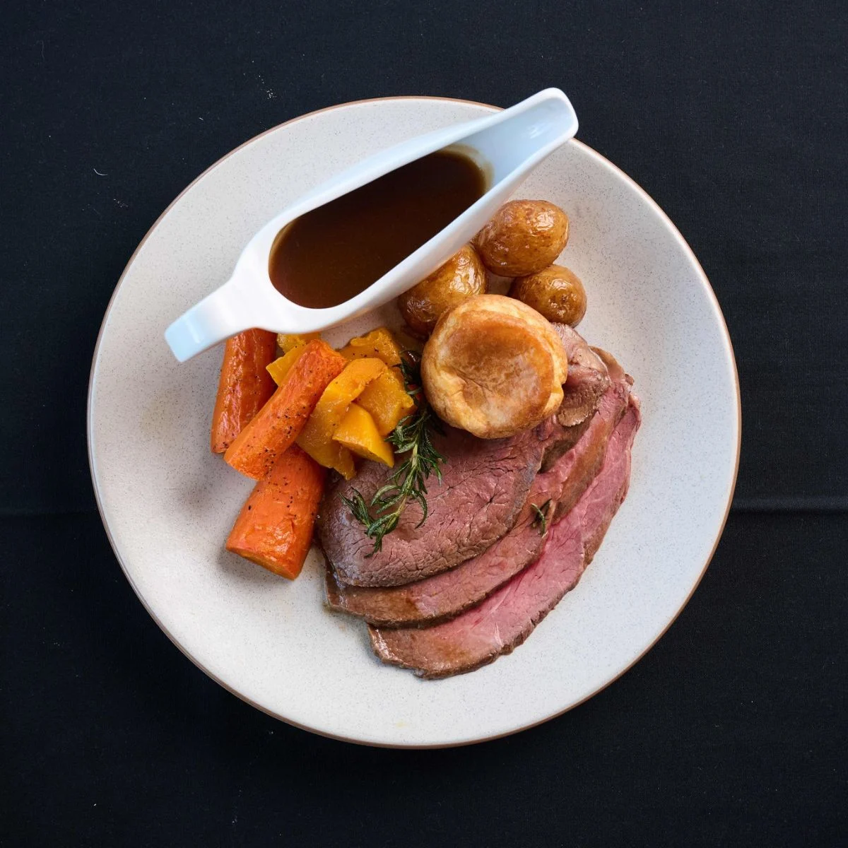 A plate of sliced roast beef with roasted potatoes, carrots, pumpkin, and a Yorkshire pudding, served with gravy in a gravy boat.