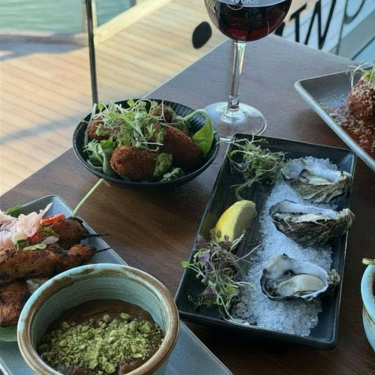 A spread of appetizers including oysters on ice, croquettes, and skewers, with a glass of red wine.