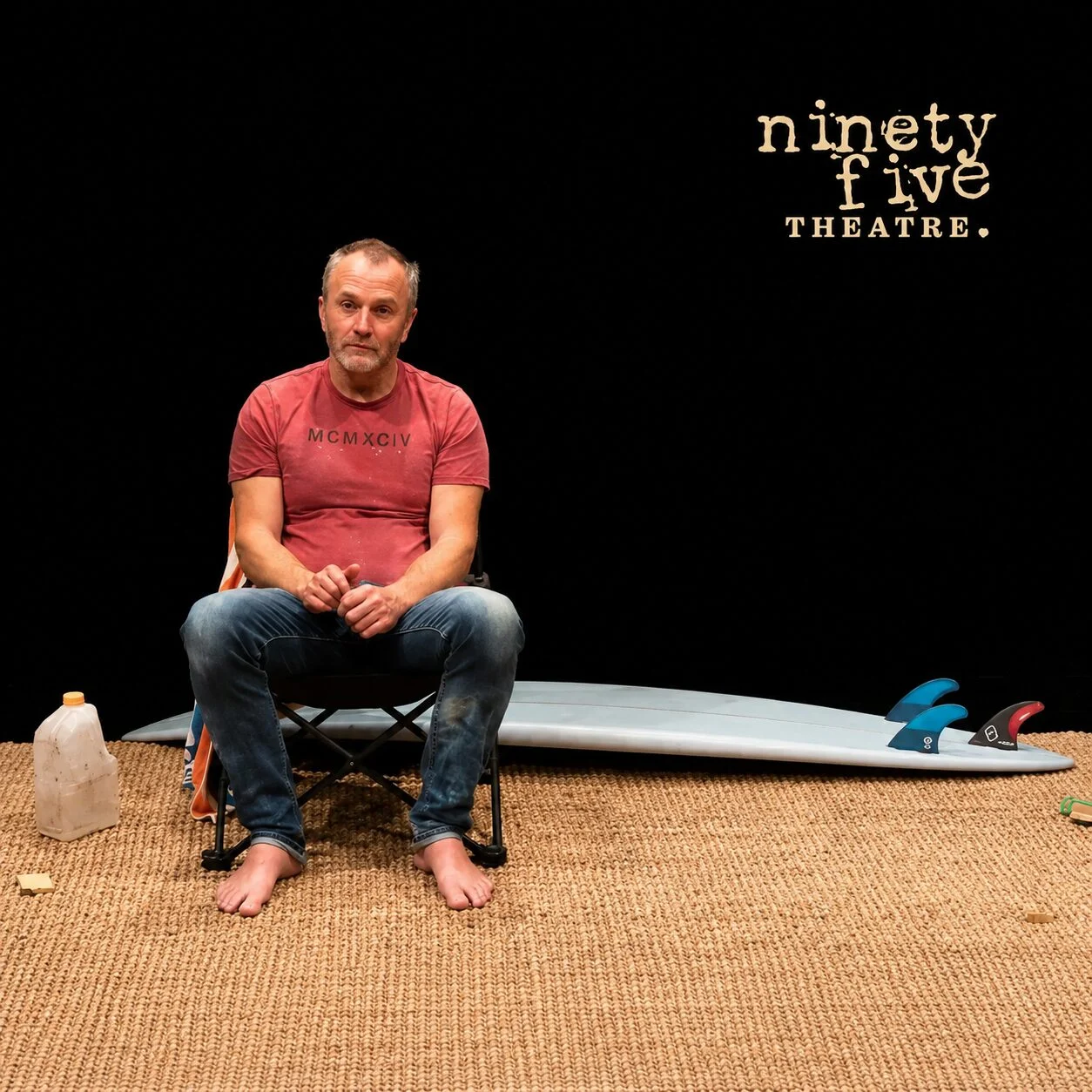 A man sits barefoot on a chair on a woven mat, with a surfboard, a plastic jug, and a cup nearby. He wears a red T-shirt and jeans. The background is black with “ninety five THEATRE.” written in the corner.