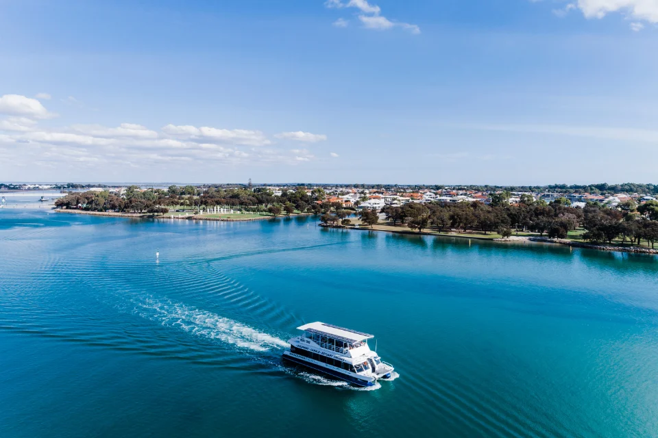 A white cruise boat moves across a calm turquoise bay, leaving a wake, with a coastal shoreline and low buildings in the distance under a clear blue sky with scattered clouds.