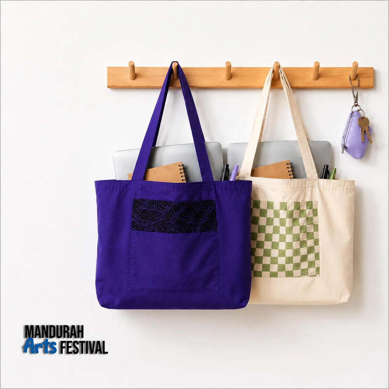 Two tote bags, one purple and one beige with a green checkered pocket, hang on a wooden rack. Each bag holds notebooks and a laptop. A small key pouch also hangs on the rack. Mandurah Arts Festival logo is at the bottom left.