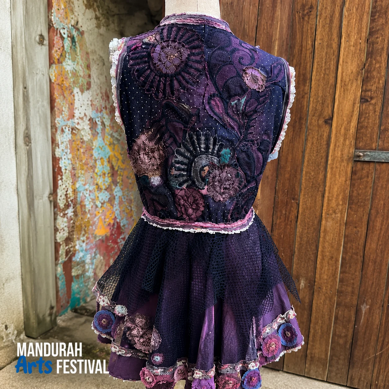 A purple, floral-embroidered dress on a mannequin stands near a rustic wooden door and a peeling, colorful wall. MANDURAH Arts FESTIVAL is written in the bottom left corner.