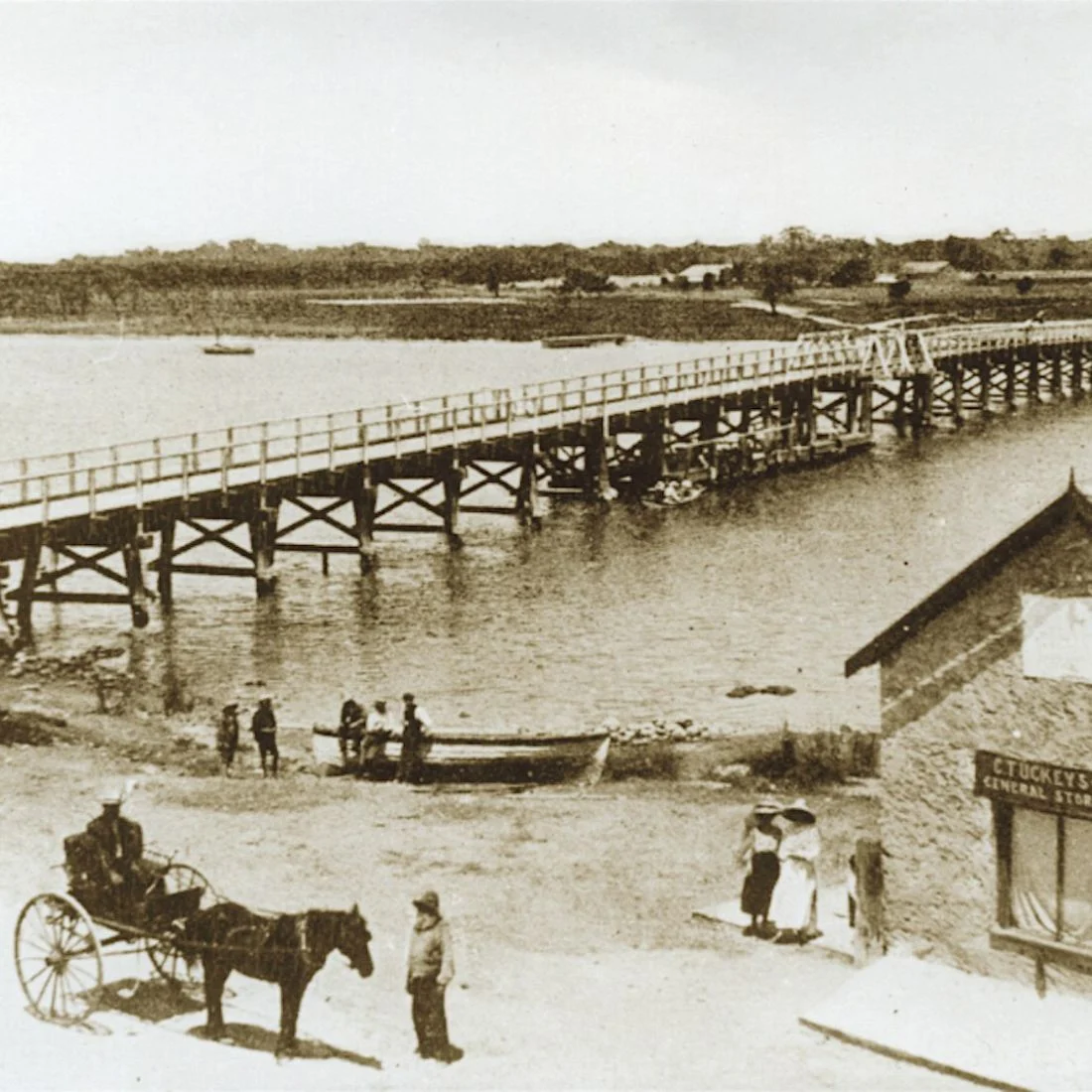 Sepia scene of a long wooden pier with railings and small boats on calm water, people walking along the shore, and a horse-drawn carriage near a roadside sign.