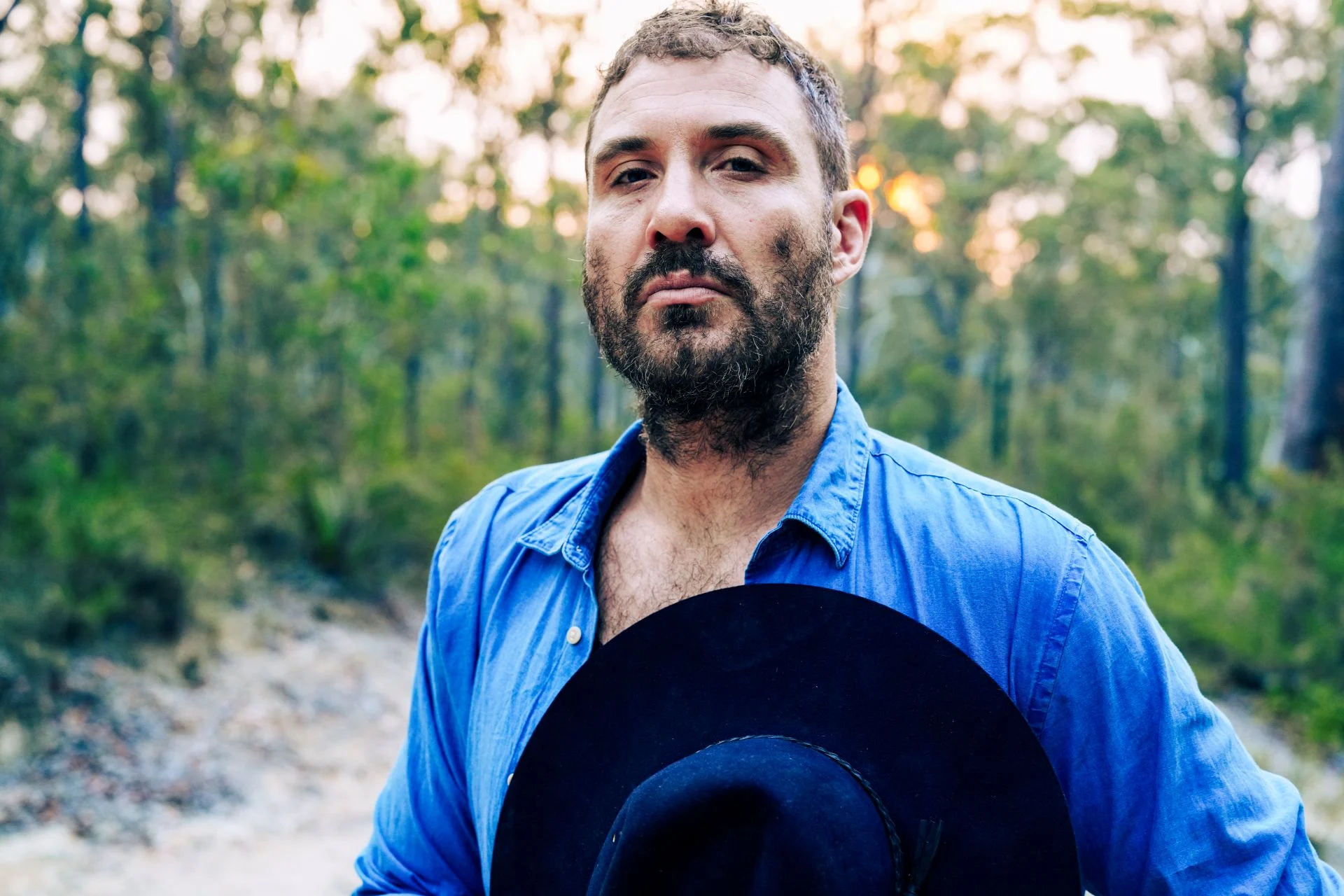 A man with short hair and a beard stands outdoors holding a black hat, wearing a blue button-up shirt, with trees and sunlight in the background.