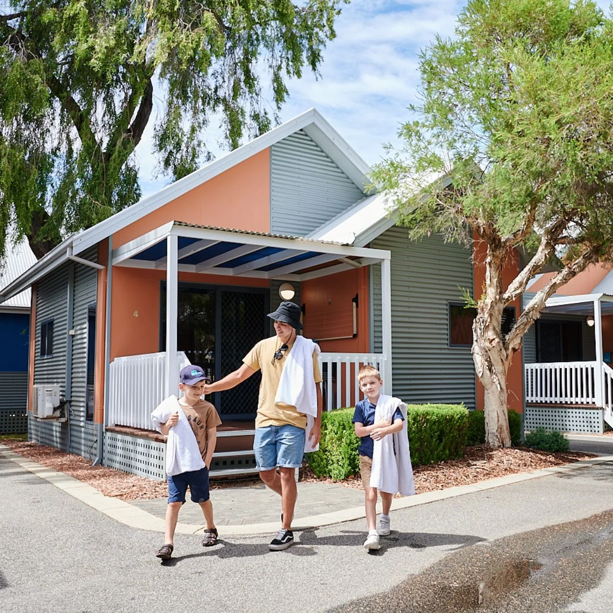 A man and two young boys walk together outside a modern cottage with corrugated siding. The man has a towel around his neck, and the kids wear towels and hats. Trees and a shaded patio are visible.