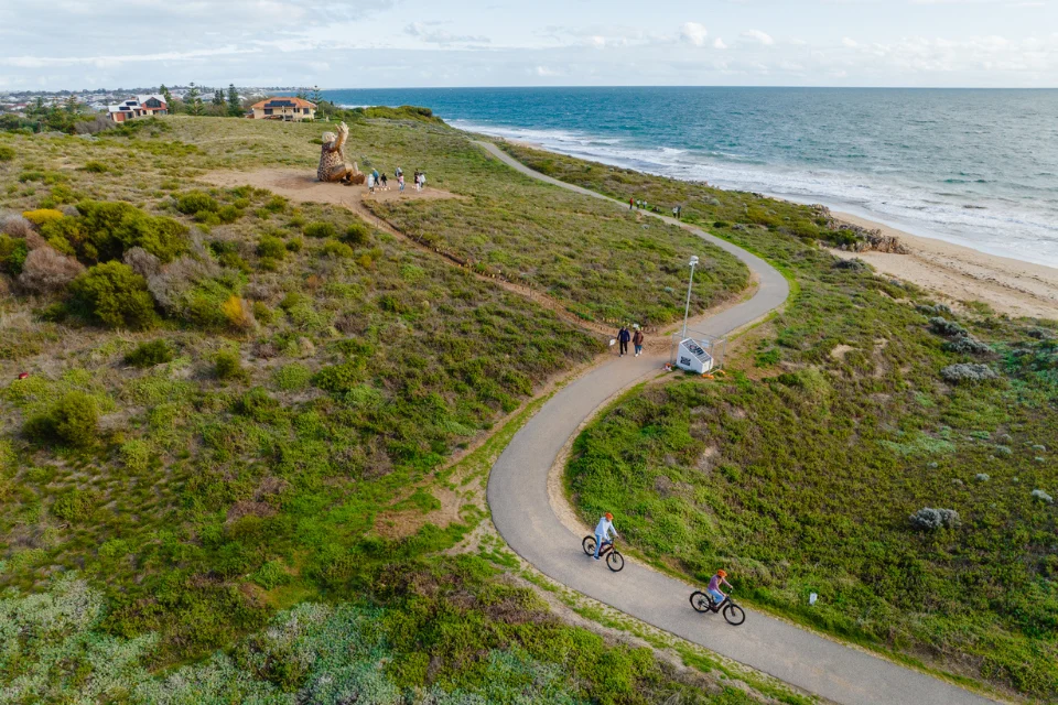 Aerial view of a winding coastal path through green dunes beside the ocean, with cyclists and pedestrians riding along the road and a small van parked near the beach.