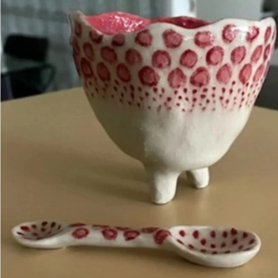 A handmade ceramic cup with three short legs and red dot patterns, paired with a matching ceramic spoon, both resting on a beige surface.