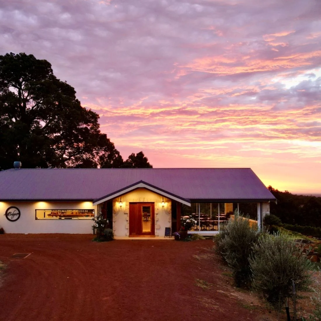 A single-story house with a metal roof sits on red soil, surrounded by trees, under a colorful sunset sky with pink, purple, and orange clouds. Warm lights glow from the houses windows and entrance.