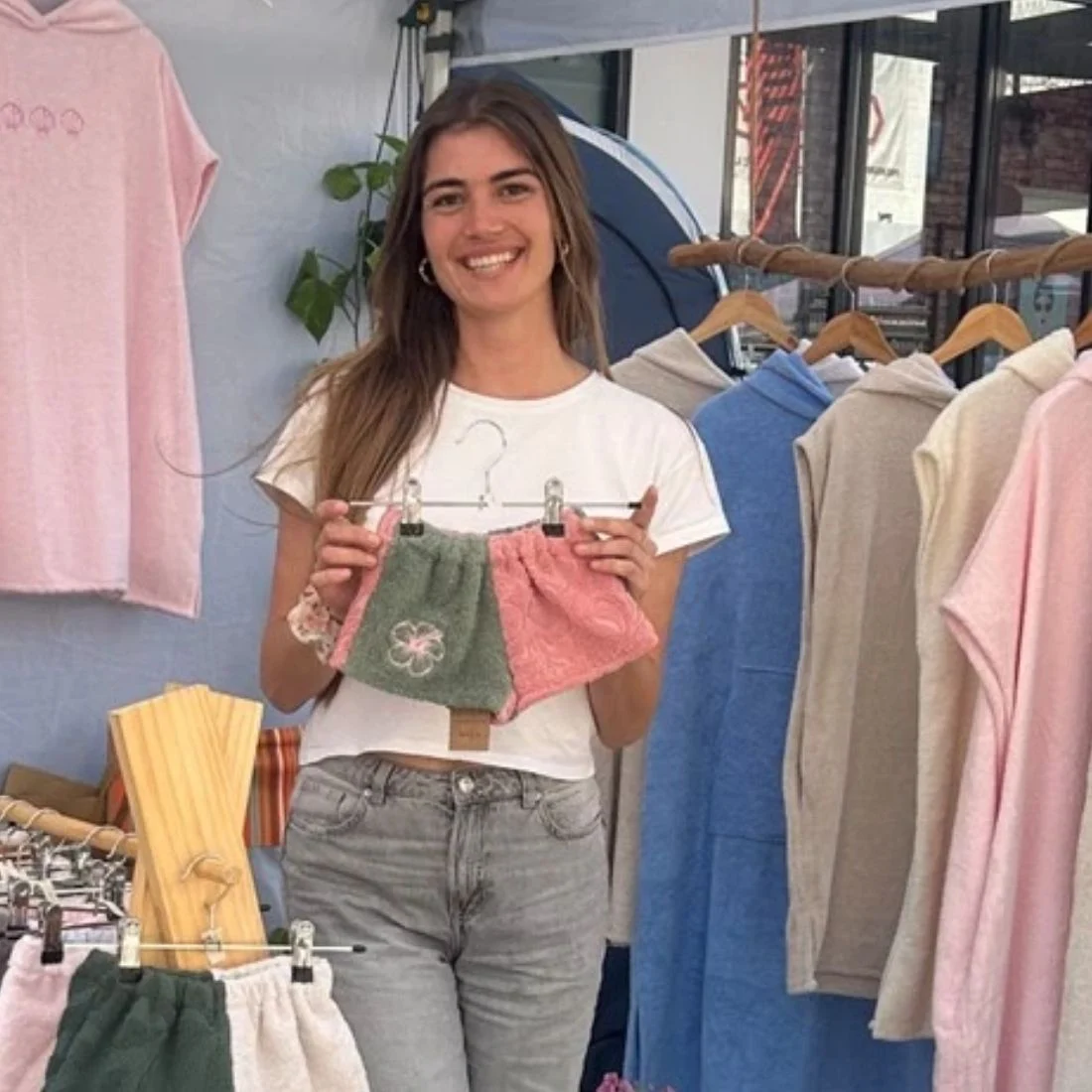 A woman stands at a clothing stall, smiling and holding up two pairs of textured shorts in green and pink. Various garments hang on racks around her.