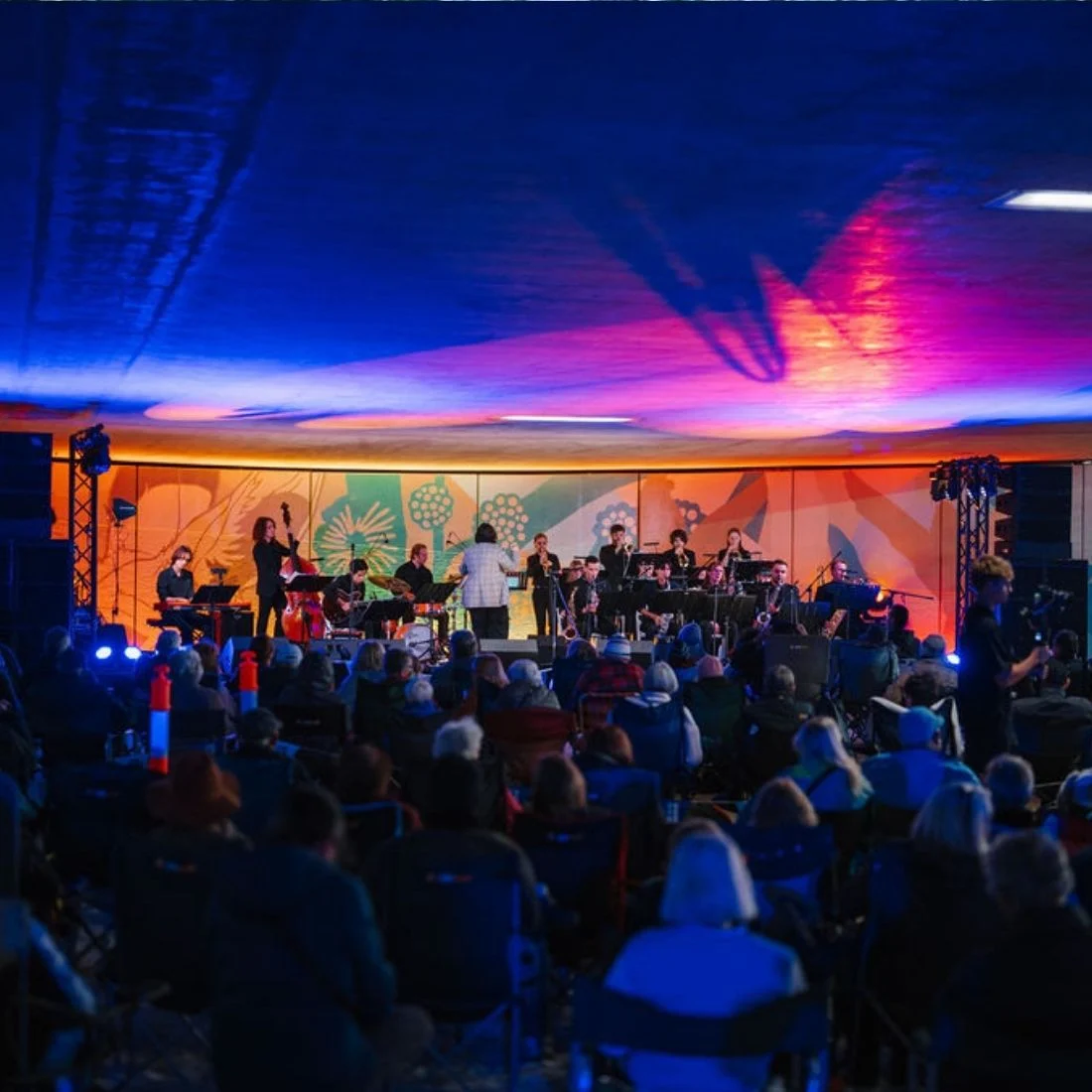 A band performs on a brightly lit stage with colorful background art, while an audience sits in chairs watching under a ceiling with blue and pink lighting.