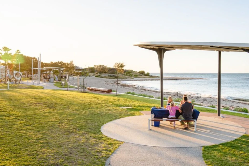 A seaside park path with trimmed grass leading to a small covered seating area; two people sit on benches as waves roll in under a clear sky.
