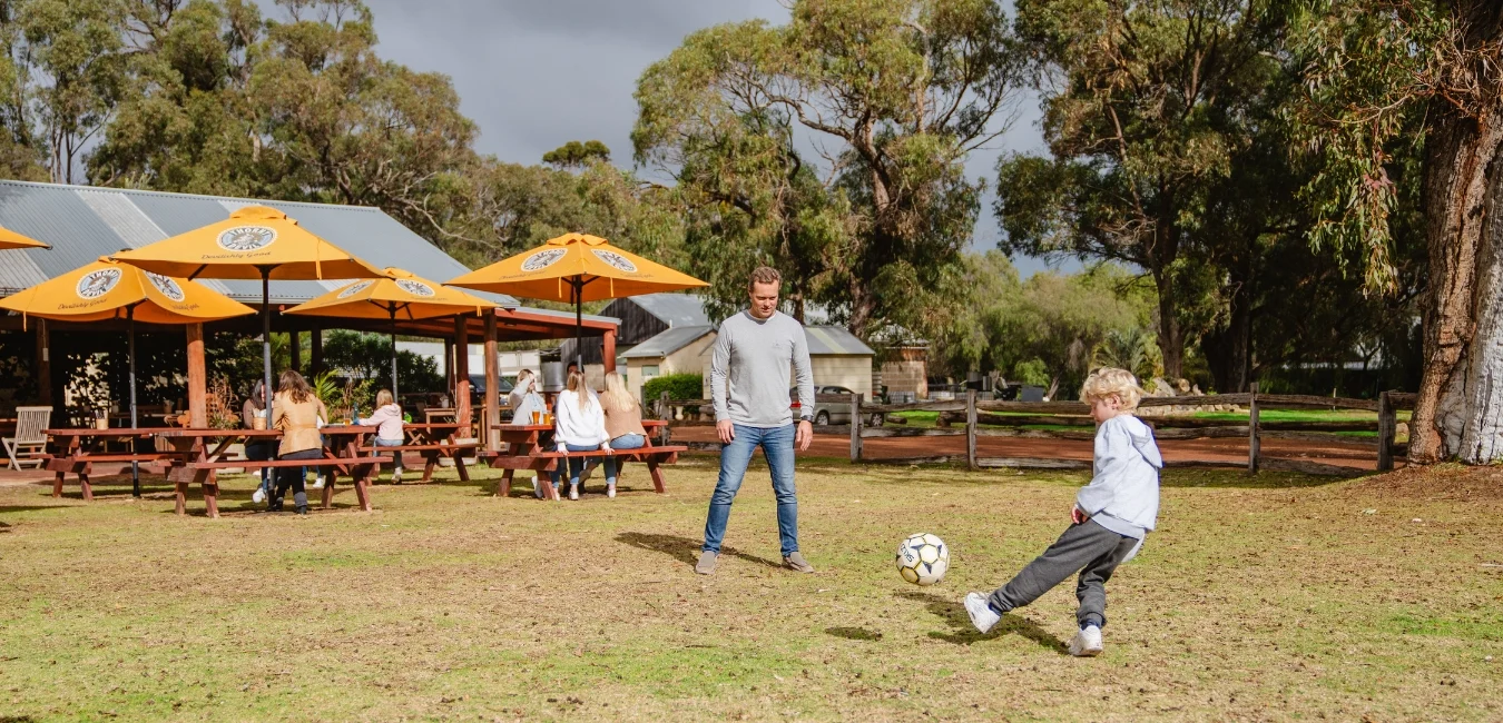 A man and a child play soccer on grass while people sit at picnic tables under umbrellas in the background.
