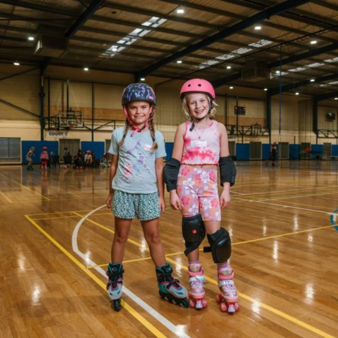 Two young girls wearing helmets and roller skates stand and smile inside a brightly lit indoor sports hall, with basketball courts and other people visible in the background.
