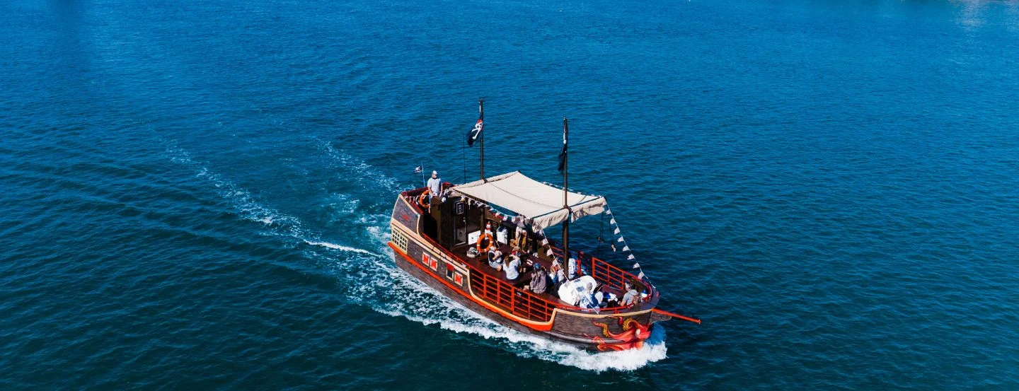 A wooden pirate-themed boat with red trim and a canopy sails on blue water, carrying several people. The boat has a dragon figurehead and creates a white wake behind it.