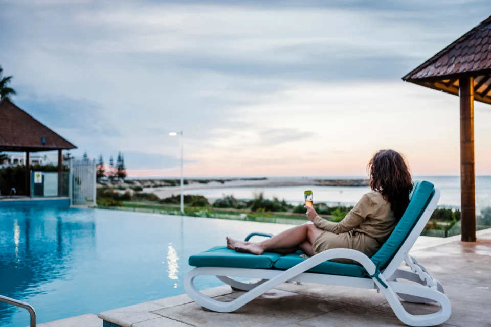 A woman with brown long hair relaxing on a blue-and-white lounge chair by an outdoor swimming pool at sunset, holding a drink, with a calm sea and distant shoreline in the background.
