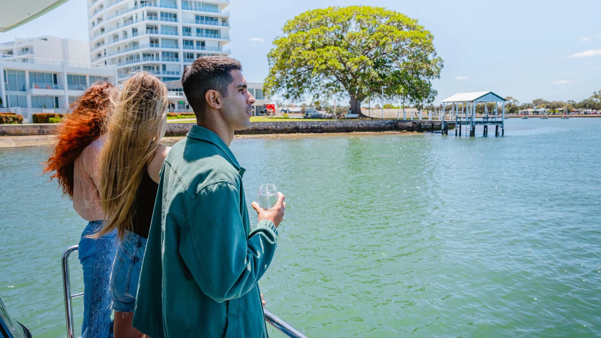 Two young women and a young man on a boat deck, looking toward the water, with a large leafy tree and modern waterfront buildings in the background.